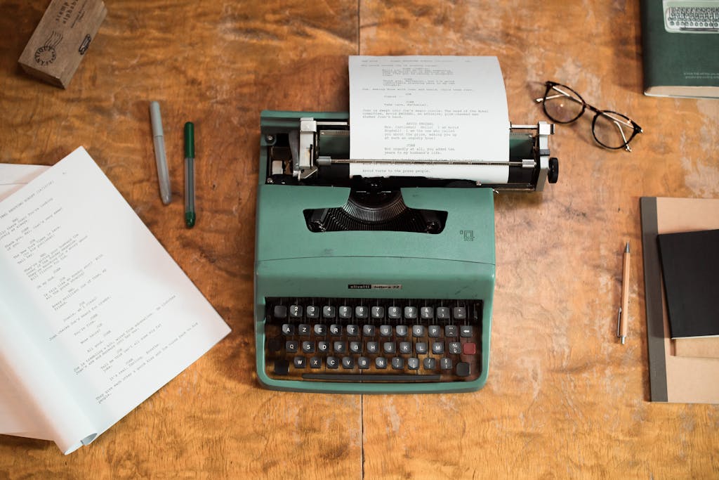 A vintage green typewriter with scripts surrounded by glasses and pens on a wooden table.